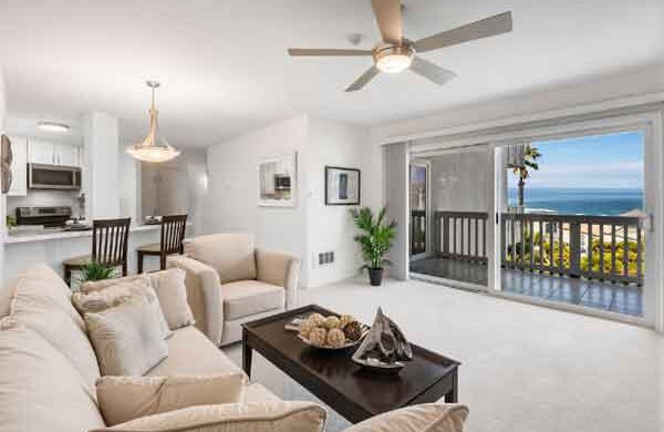 Bright open-concept living room with a beige sectional, coffee table, and sliding glass doors to a balcony with an ocean view beneath a ceiling fan.