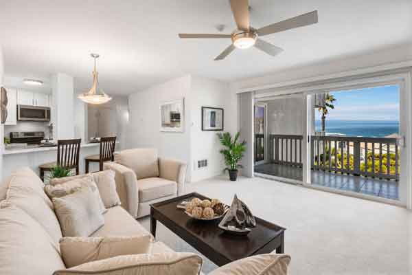 Bright open-concept living room with a beige sectional, coffee table, and sliding glass doors to a balcony with an ocean view beneath a ceiling fan.