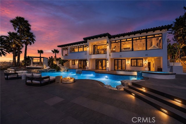 Two-story white villa with large glass walls, illuminated interiors, and a curved blue pool/spa area at sunset, palm trees nearby.